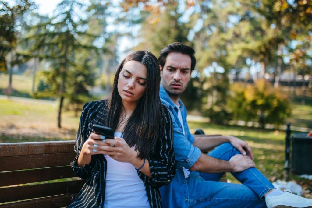 couple sitting on park bench woman on phone Signs of Controlling Men