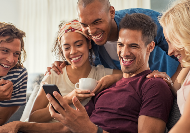 group sitting behind man with phone smiling 251 Best Paranoia Questions Game To Play With Friends